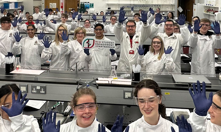 Students and staff hold gloved hands aloft in lab setting
