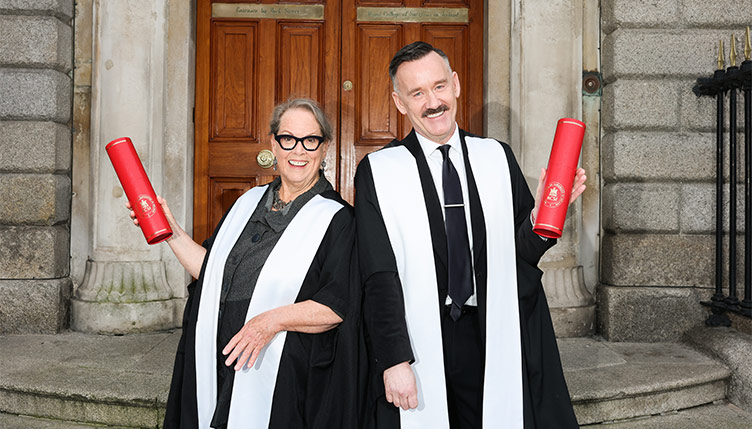 Two individuals in academic robes standing outside the RCSI building, each holding a red ceremonial scroll during a recognition or conferring ceremony.
