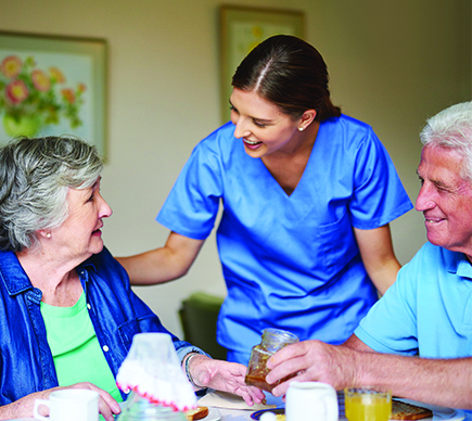 A nurse checking in on two elderly people while they eat a meal.