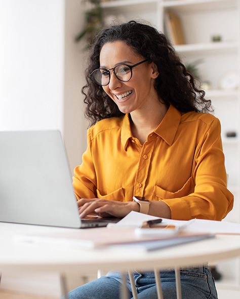 A woman wearing glasses and an orange top sitting at a desk and working on a laptop.